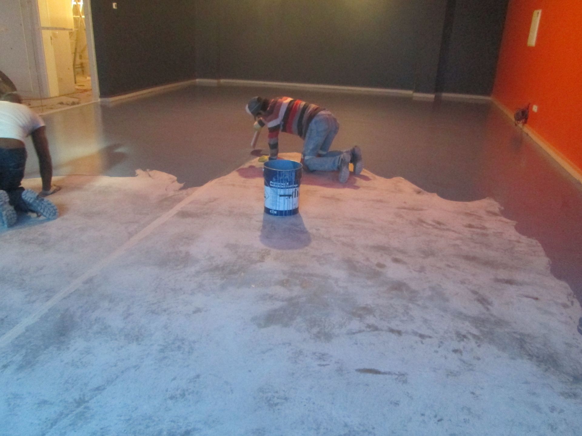 Two workers applying a coating to a concrete floor in a room with orange and dark walls.